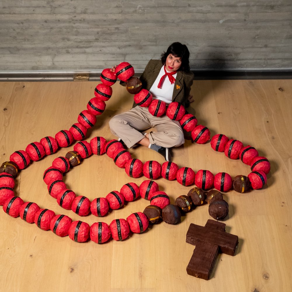 Artist Diana Benavidez sitting on the ground with a large 10 foot red piñata rosary on her lap and the floor in front of her.