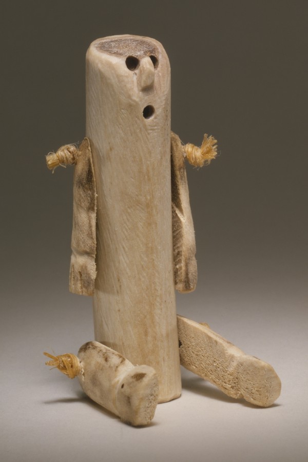 This Inuit Doll is made from a caribou antler. The body of the doll is cylindrical. Two arms and two legs are attached to body. The arms are positioned straight down to the ground. The legs are positioned out in front as if the doll was seated. The top of the caribou antler has been rounded to create a face. The mouth and eyes are open holes, while the nose was carved..