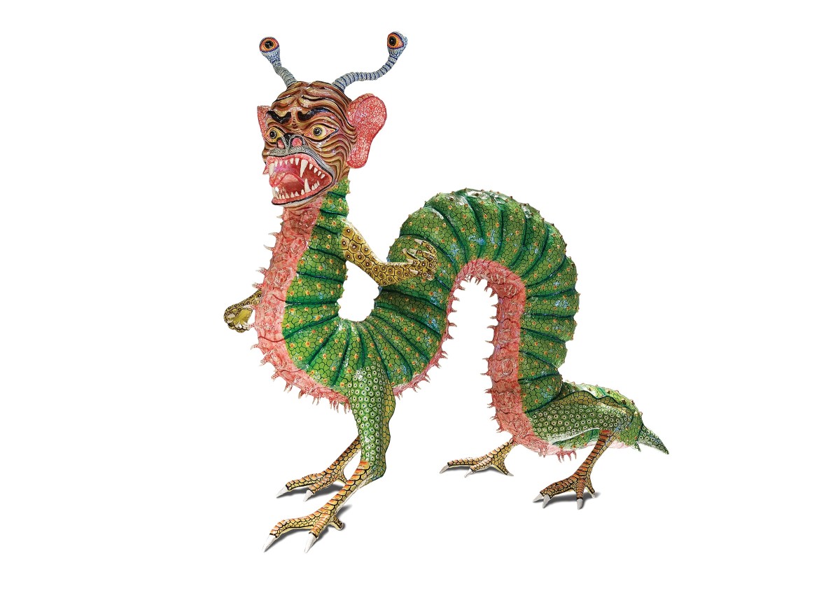 An Alebrije is a brightly colored Mexican Folk Art sculpture typically depicting a mythical creature. This Alebrije has the body of a caterpillar, 4 webbed feet and a creature-like face. It’s green with yellow spots and its underbelly is pink with red veins spidering out. It's has large, human-like ears, an antennae on top of its head. Its face has large expressive eyes, with stripes on its face and its mouth is open exposing its sharp teeth.