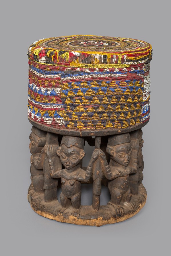 This stool was carved from a single block of wood. Its openwork base features caryatid figures (female figures, used as a pillar to support the entablature of a Greek or Greek-style building). The top of the stool is decorated with beads which indicates it was intended more for display at public events and ceremonial occasions rather than for everyday seating.