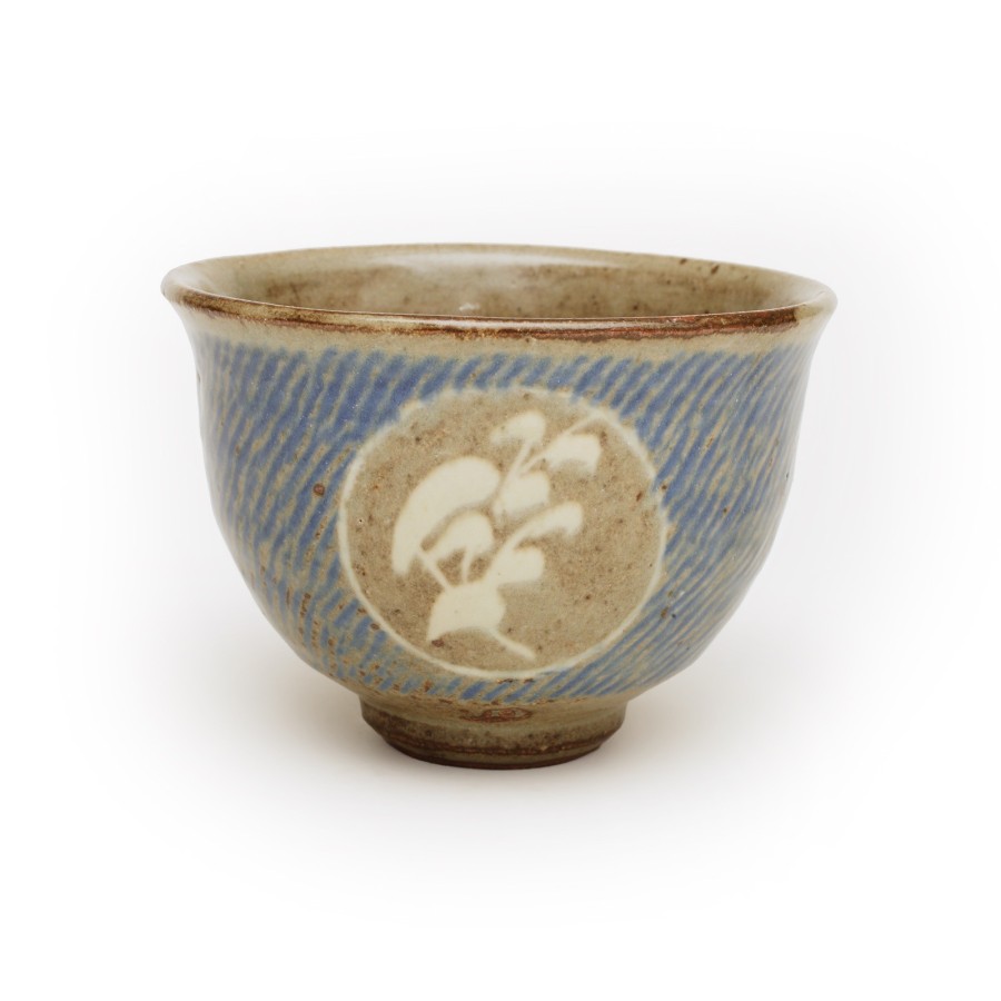 This stoneware tea bowl is brown with blue glaze over a diagonal texture design. The textured pattern was made by impressing rope into the damp clay prior to firing in the kiln. There is also an untextured circle on one side the bowl that exposes the stoneware overlaid with a white simplistic plant motif..