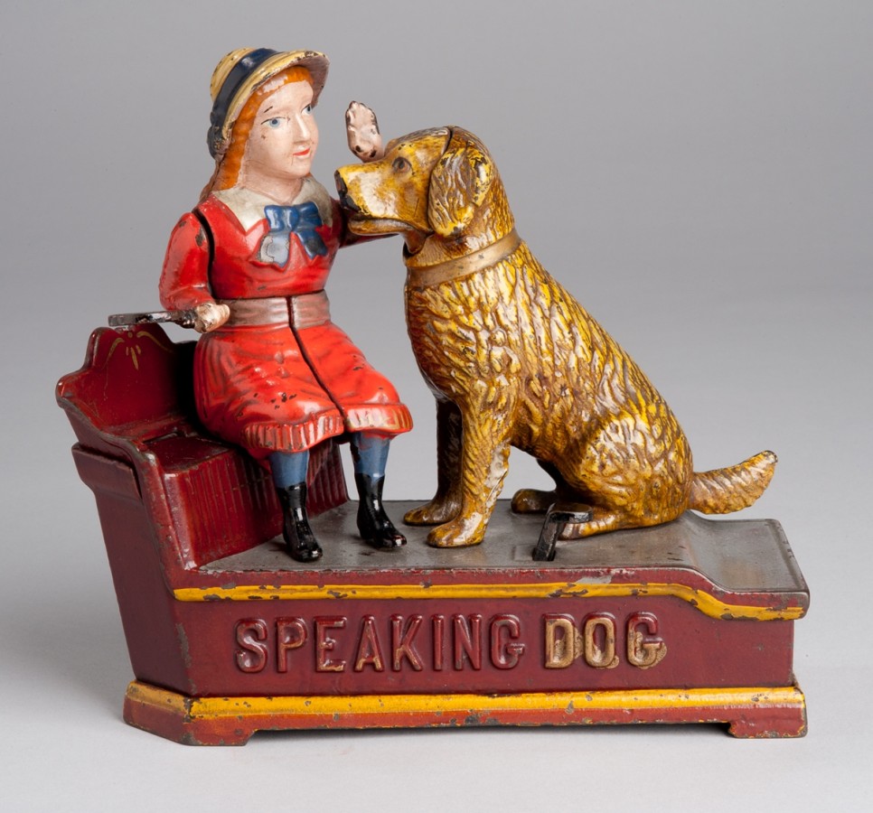 This cast iron mechanical toy bank features a young girl sitting on the bench petting her dog. The dog is in a seated position and is the same height as the girl sitting on the bench. The girl is wearing a read dress with a blue bow and possibly a straw hat. Underneath the girl and the dog are the words "speaking dog".