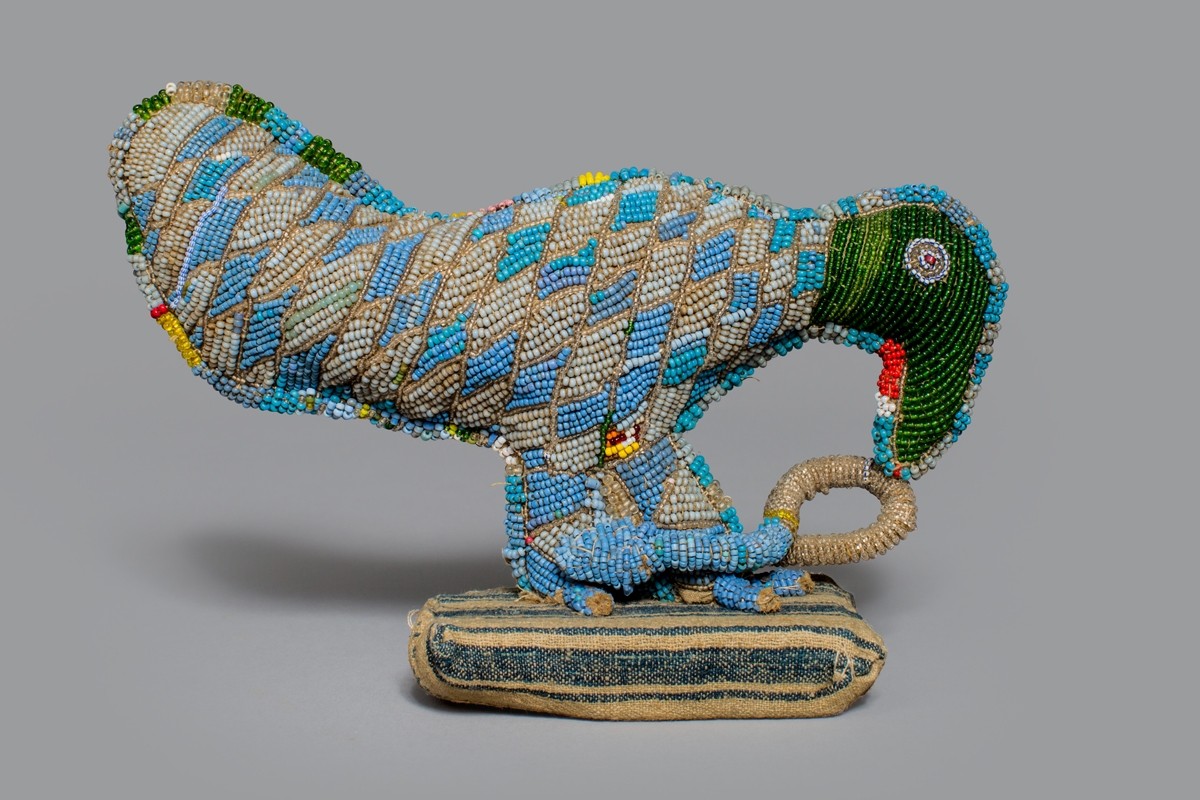 Only seven inches tall, this figure comprised of glass beads, cotton, and wood depicts a bird attacking a snake with its beak. The body of the bird is beaded with blue, green, and cream beads in a checkered diamond pattern. The head of the bird uses green beads. The snake is white beads.