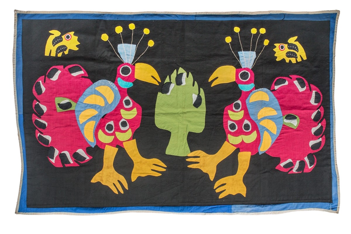 This textile is an appliquéd and sewn cotton banner typical of the Fon people of Benin. The textile is longer horizontally than vertically. The background is black and there are two large pink and blue stylized peacocks. The two peacocks cover the majority of the textile and are facing each other. In the upper right and left corner is one small yellow creature, possible a cat. In the center of the textile, in between the two peacocks is a large green motif, similar in shape to an artichoke.